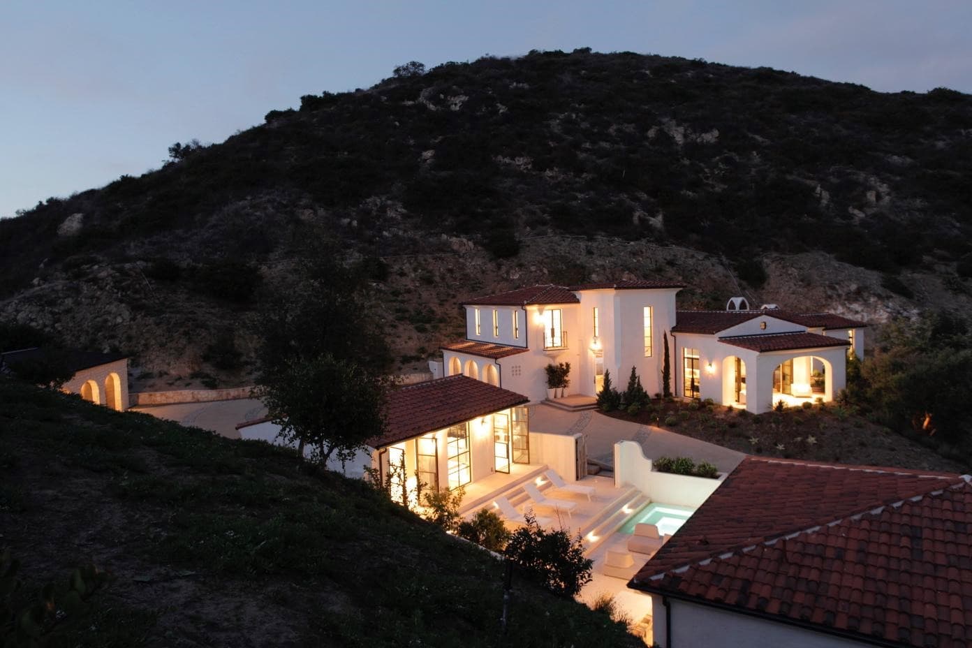 Hillside Spanish-style home at dusk with white stucco walls, red tile roofs and lit windows