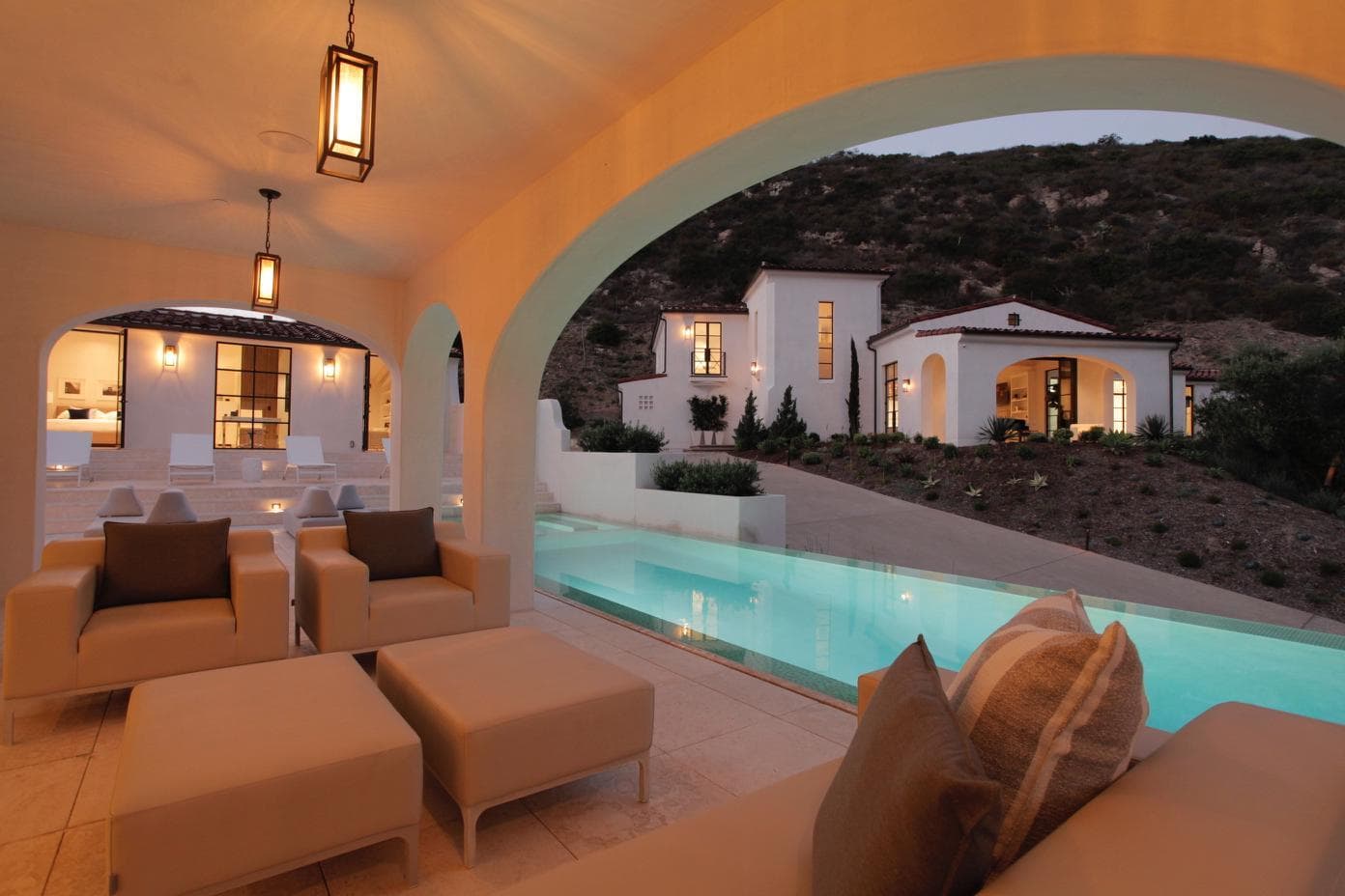 Covered terrace at dusk with pendant lanterns, looking across the pool to the main house
